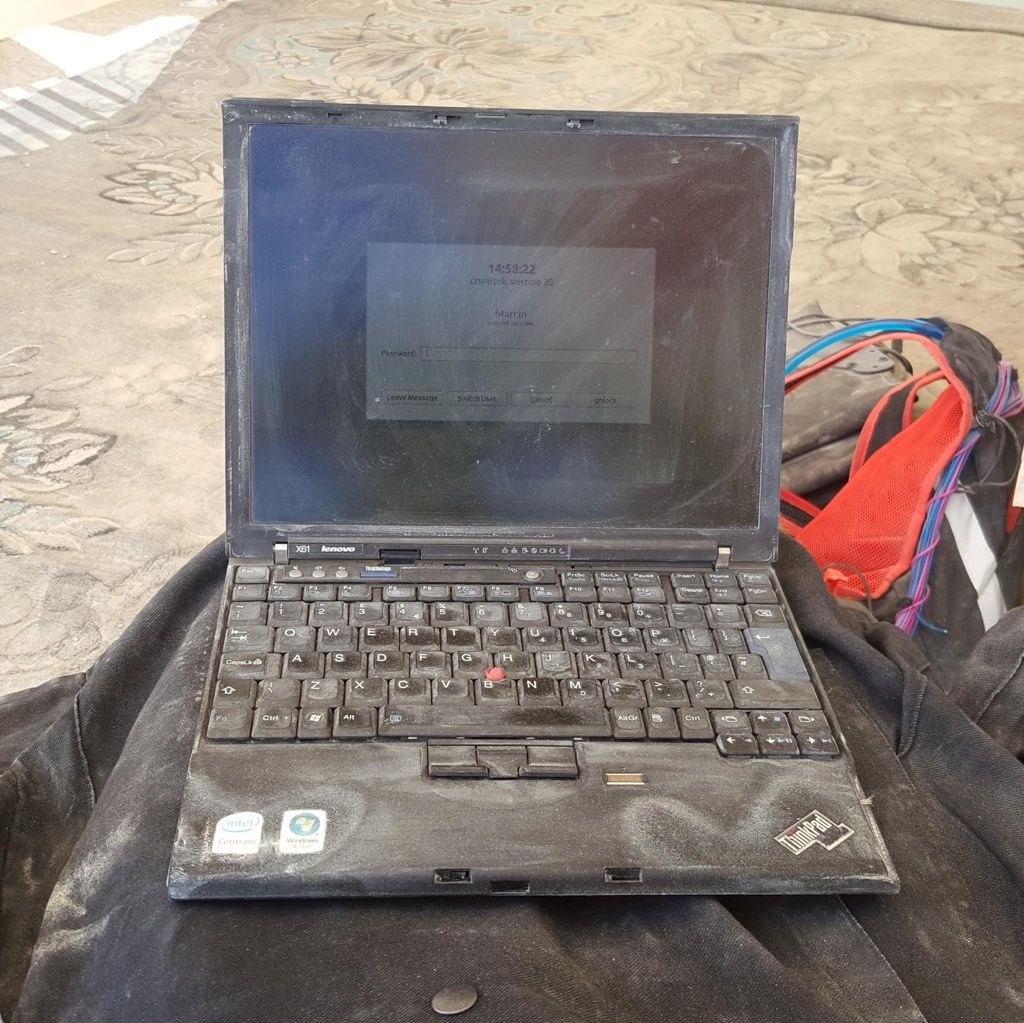 An old ThinkPad x61, back from the 4:3 era, completely dusted over but open and working.  A backpack on a dirty and dusty carpet in the background.