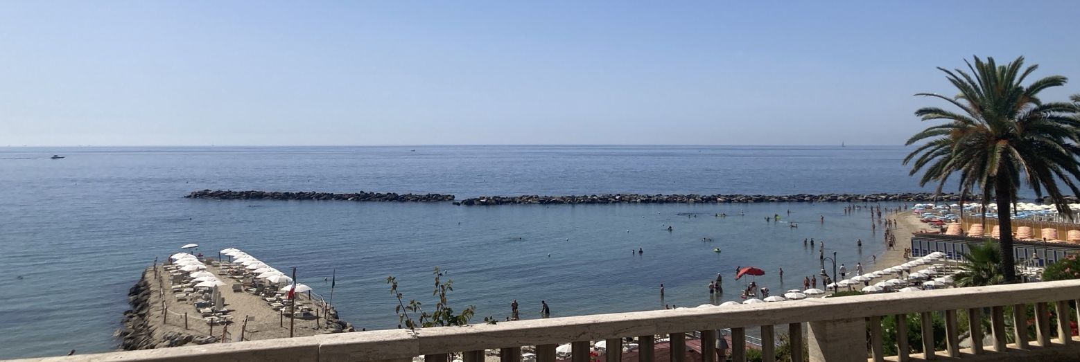 Clear blue sky over clear blue water with some people, lots of beach umbrellas, a molo and a breakwater all visible.  A palm to the right.